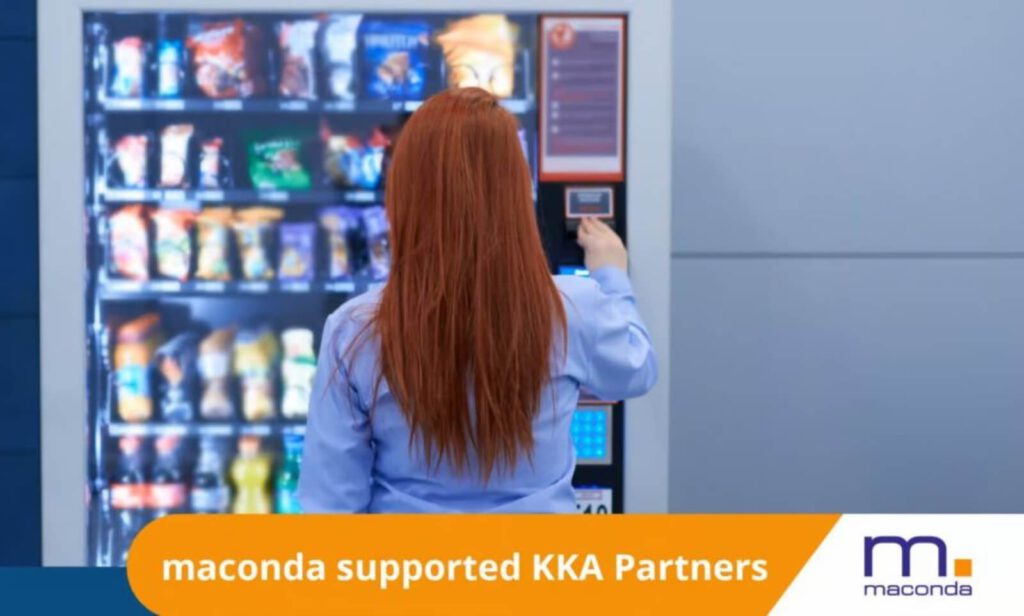 maconda News Dealbanner-KKAPartners 250301 - maconda Person with long red hair in a blue shirt operating a snack vending machine seen from behind orange banner maconda supported KKA Partners