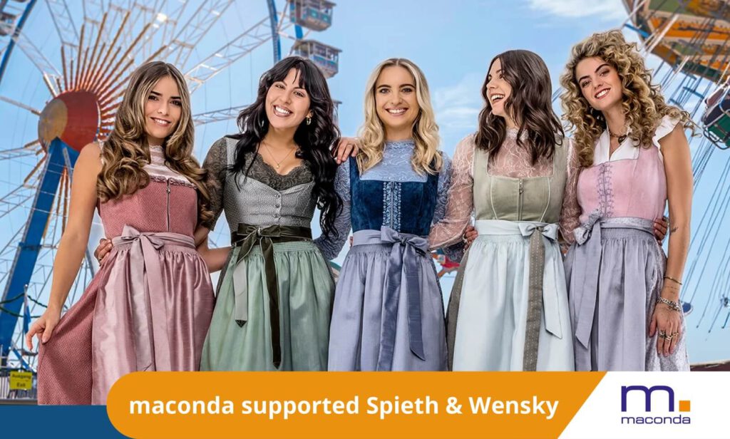 Five women in colorful dirndls stand arm in arm in an amusement park in front of a Ferris wheel orange banner maconda supported Spieth & Wensky