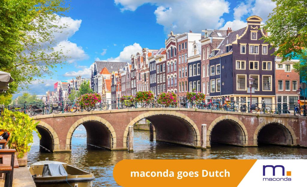 Historic canal houses on a brick bridge in Amsterdam bicycles on the railing under a blue sky banner with maconda goes Dutch