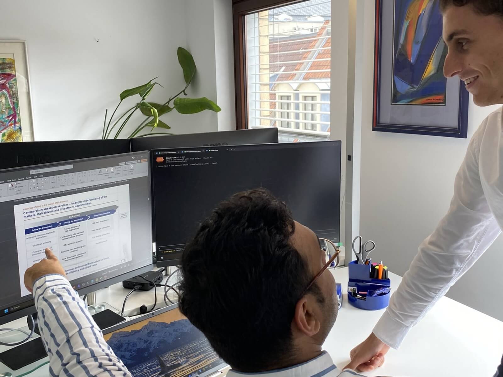 Zwei Kollegen arbeiten an einem Schreibtisch mit zwei Monitoren einer zeigt auf ein Dokument auf dem linken Bildschirm
