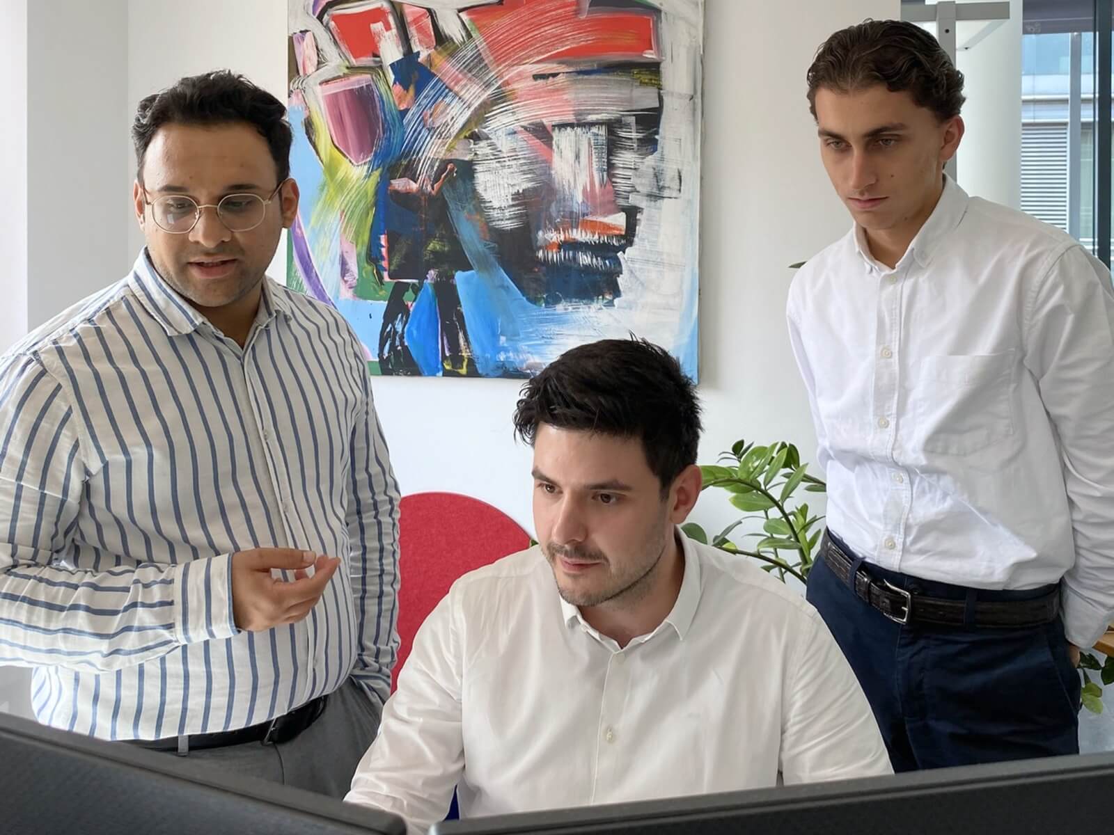 Drei Männer in Business Casual Kleidung stehen um einen Monitor in einem hellen Büro dahinter ein abstraktes Gemälde