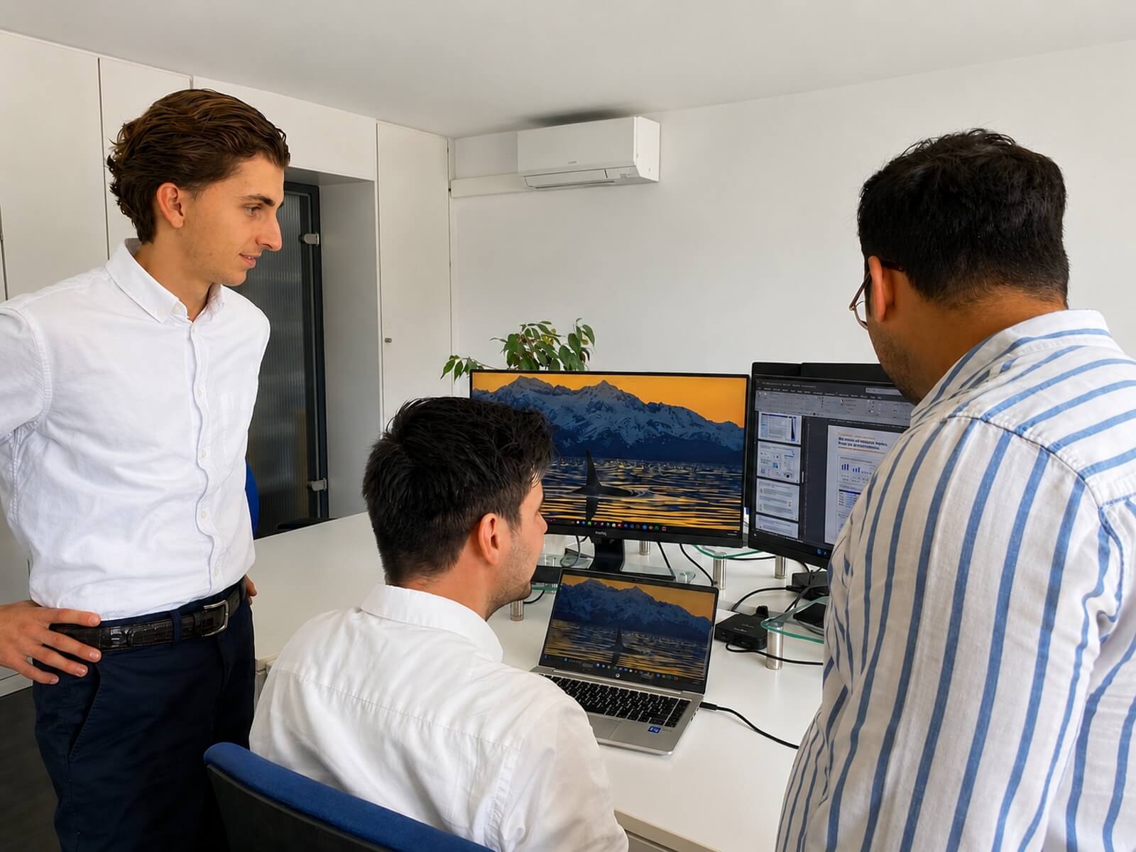 Three colleagues standing and sitting in a bright office around a white desk checking data on two monitors and a laptop