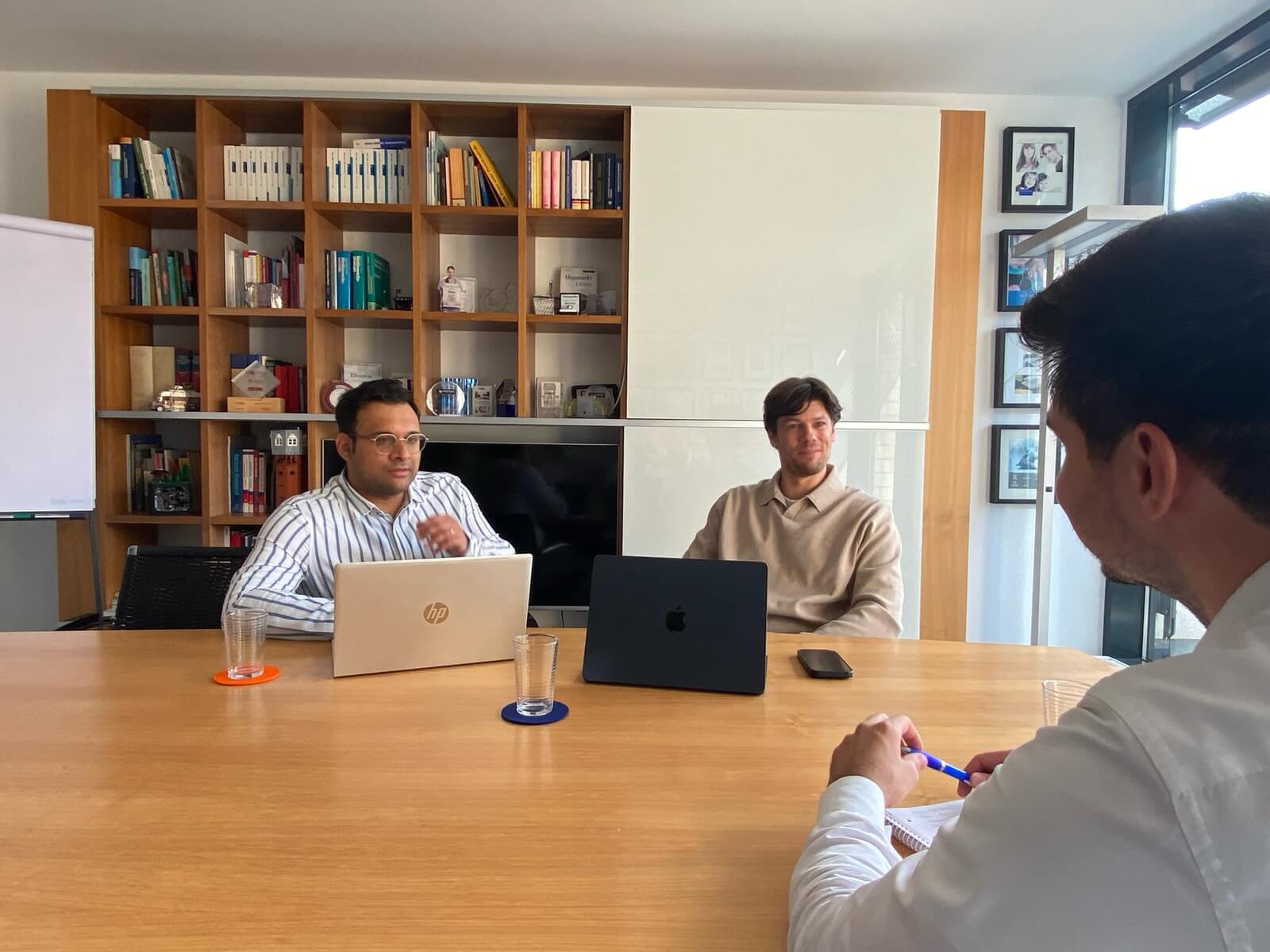 Drei Männer sitzen mit Laptops an einem Konferenztisch aus Holz in einem hellen Büro vor einem großen Bücherregal