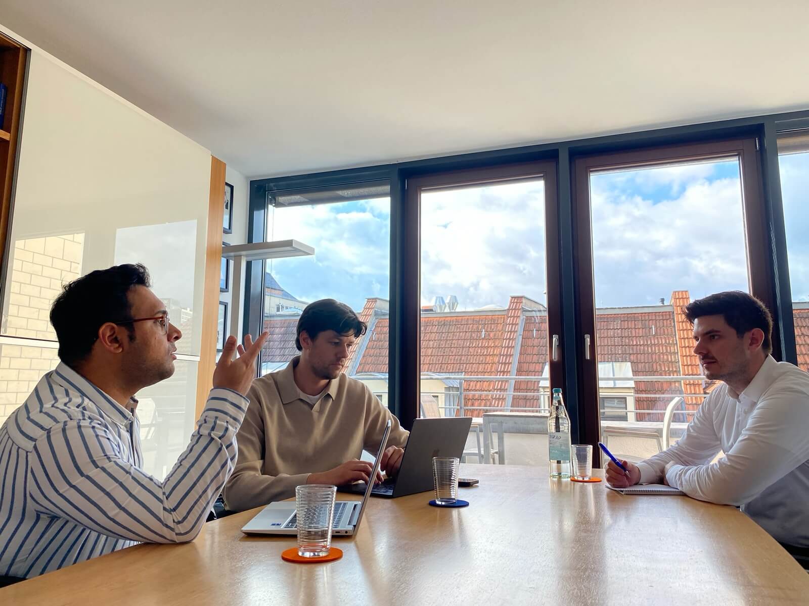 Three men sitting with open laptops at a wooden table in a bright office discussing