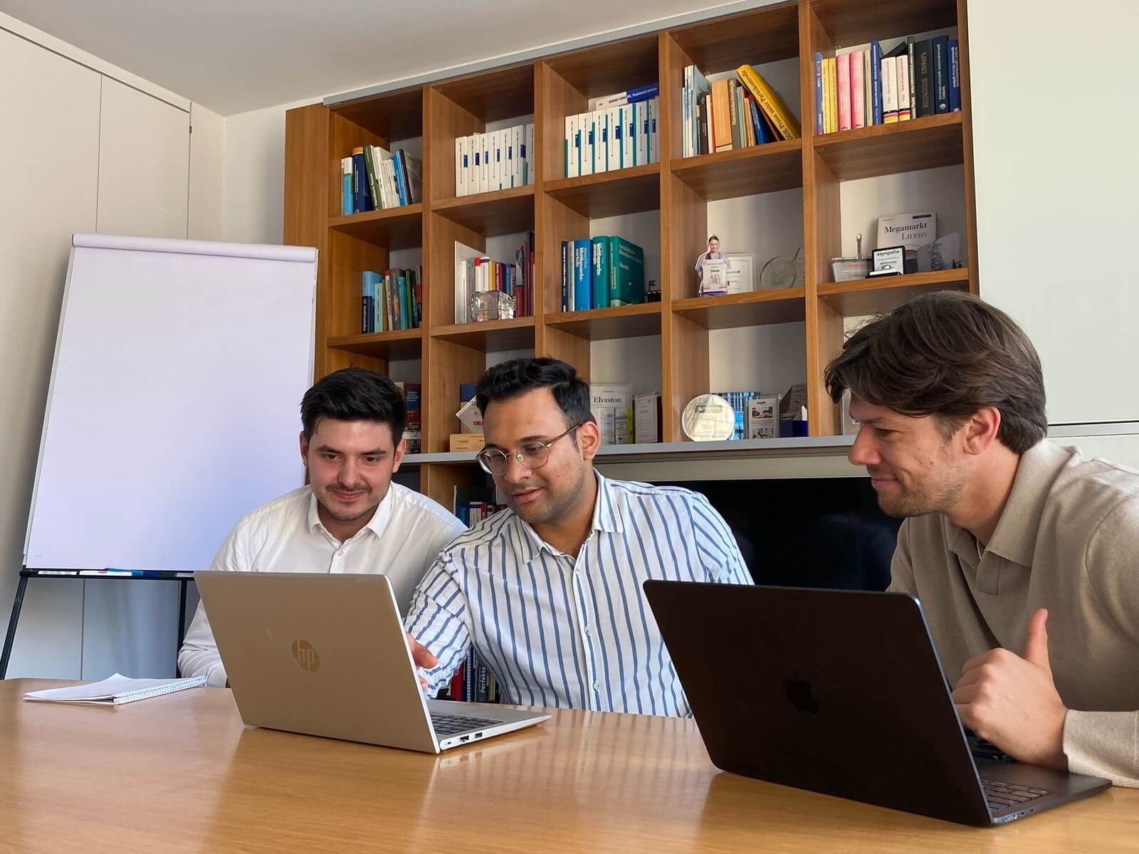 Drei Kollegen sitzen mit Laptops an einem Konferenztisch aus Holz in einem hellen Büro dahinter ein hohes Bücherregal und links ein leeres Flipchart