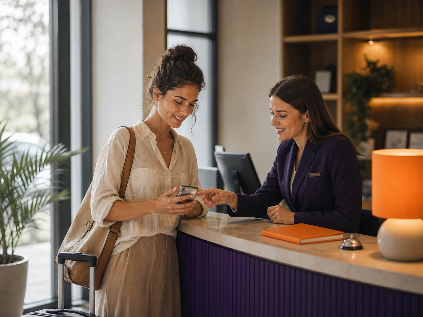 Hotel reception: Smiling guest with suitcase shows receptionist his smartphone at the counter.