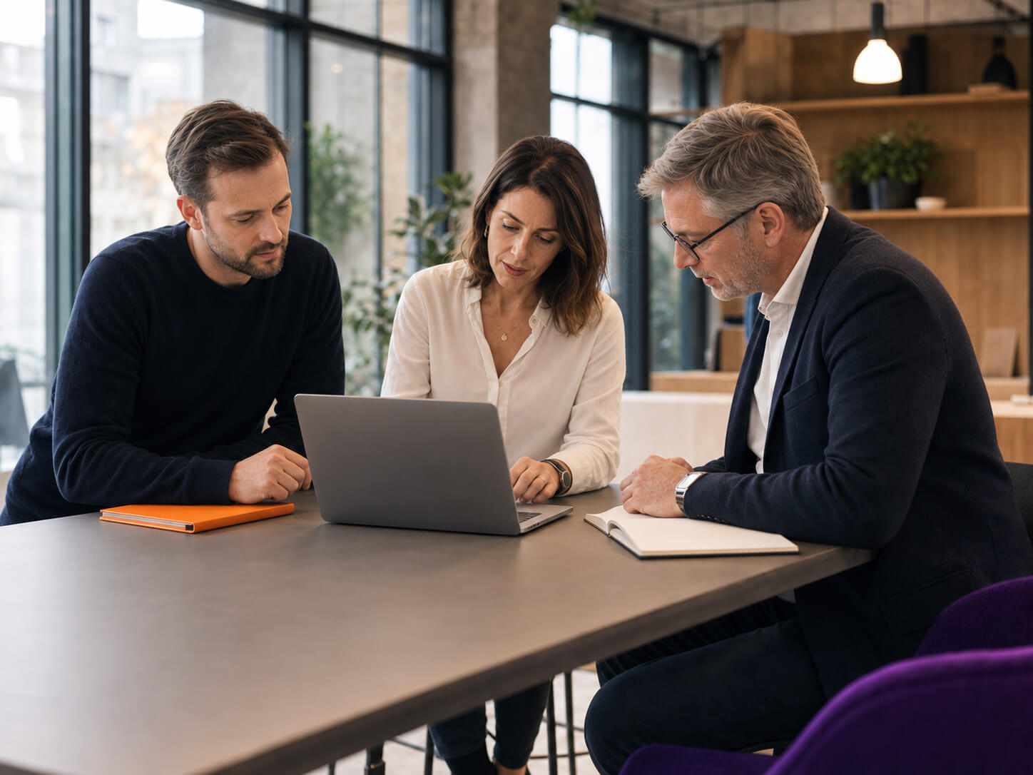 maconda-geschaeftsmodell-versteher - maconda Drei Kollegen sitzen an einem Tisch und schauen gemeinsam konzentriert auf einen Laptop in einem modernen Büro