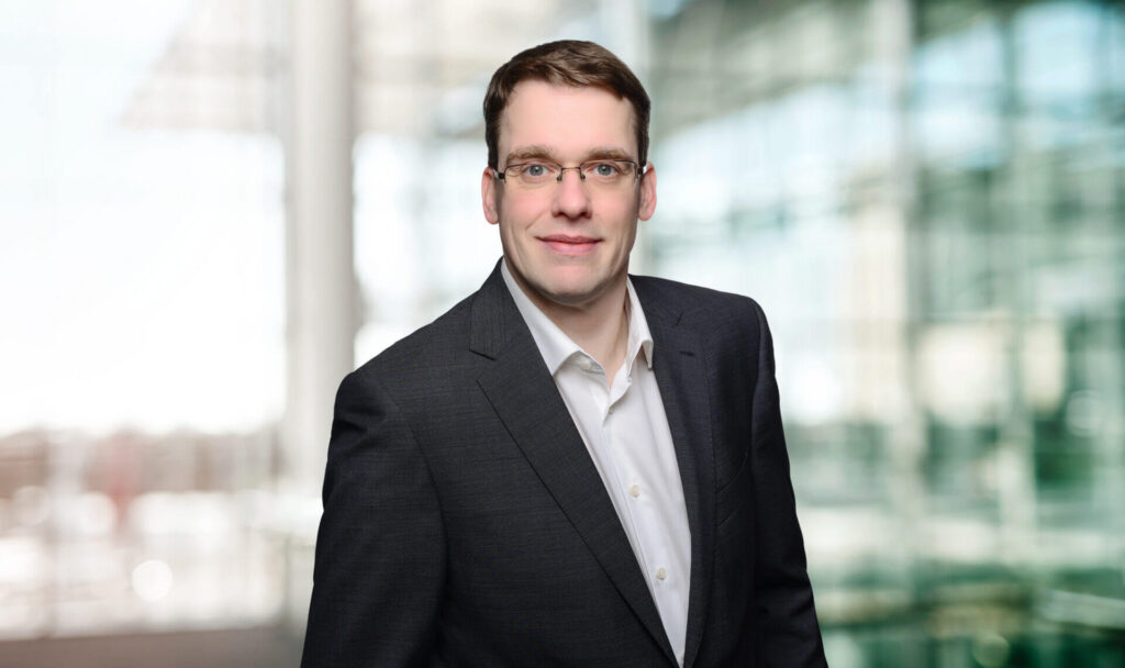Portrait of Bertram Stähler maconda in a dark suit and white shirt smiling in front of a blurred office background