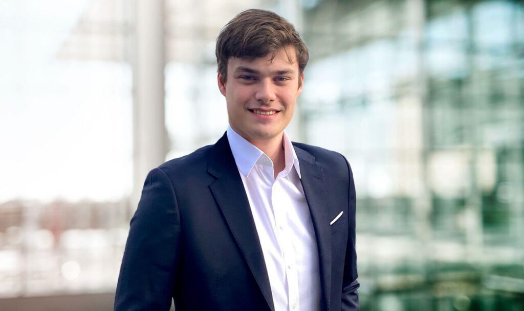 Porträt von Lukas Dedy maconda in dunkelblauem Anzug und weißem Hemd lächelnd vor einem modernen Glasgebäude