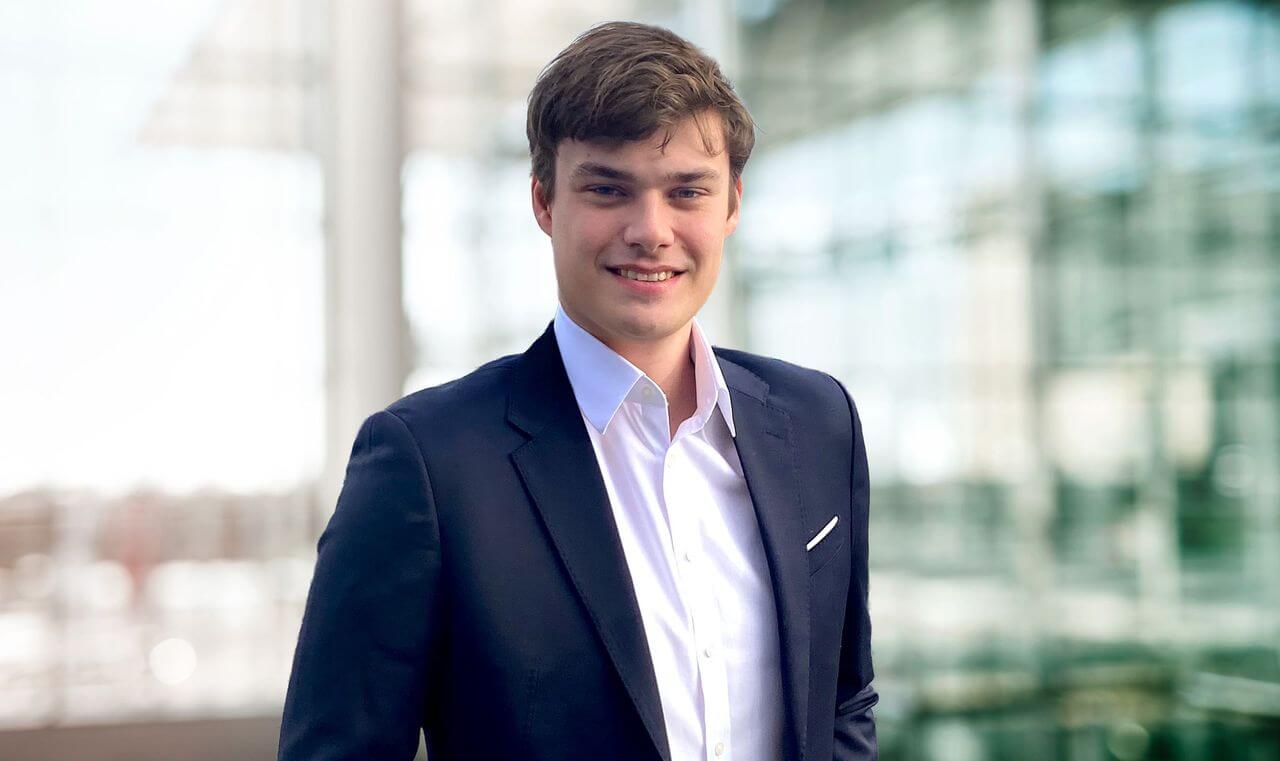 Porträt von Lukas Dedy maconda in dunkelblauem Anzug und weißem Hemd lächelnd vor einem modernen Glasgebäude