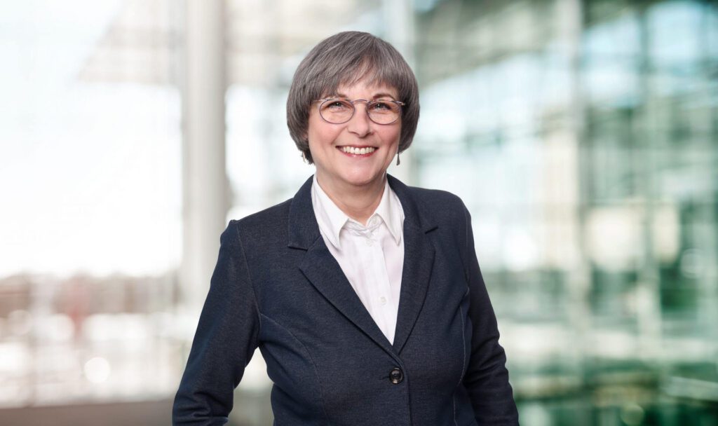 Portrait of Marion Tichel maconda in a dark blazer and white blouse smiling in front of a bright office background