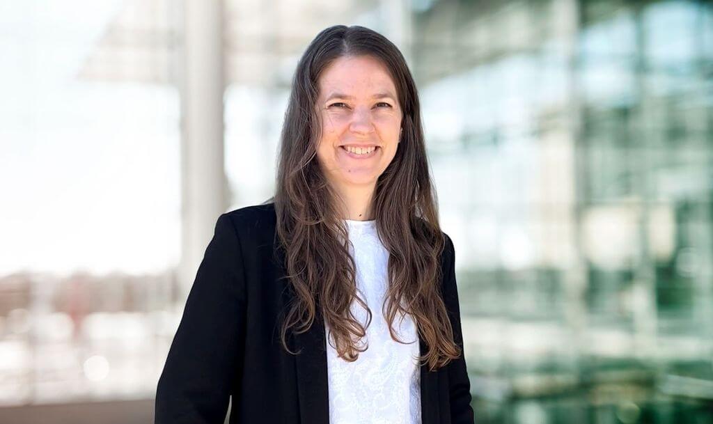Porträt von Sara Baier maconda in schwarzem Blazer und weißer Bluse lächelnd vor einem Glasgebäude